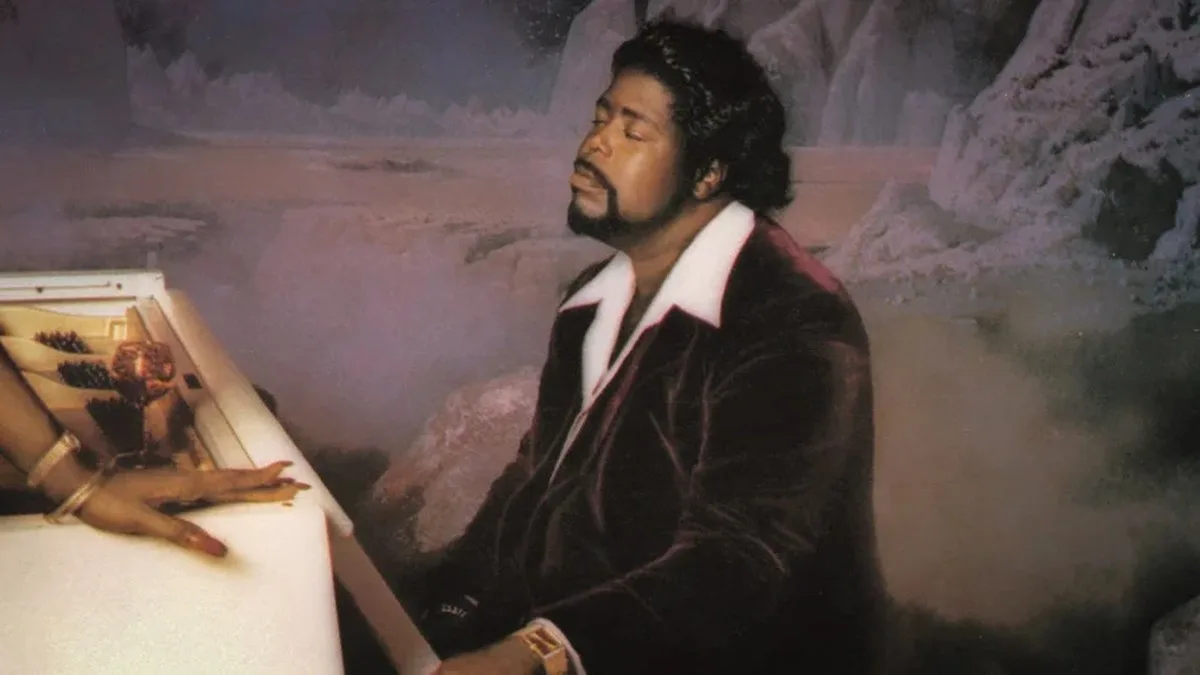 A Black man with coiffured hair and a velvet jacket and white shirt beneath it, sits eyes closed at a piano, as if dreaming or in some kind of state of pleasure. Mists swirl around him. It's singer Barry White.