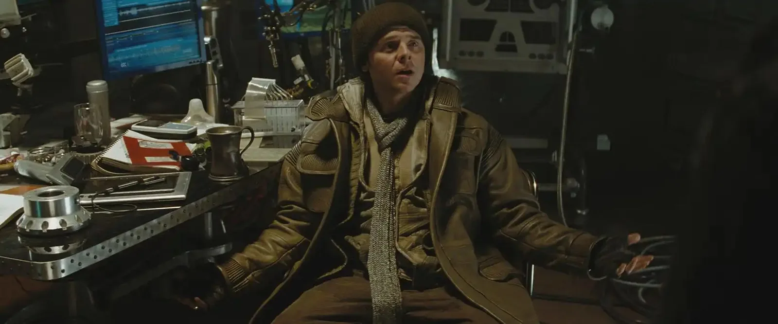 Simon Pegg as Scotty in Star Trek (2009) spreads his arms in a questioning gesture, sitting in warm clothing at a cluttered desk. 